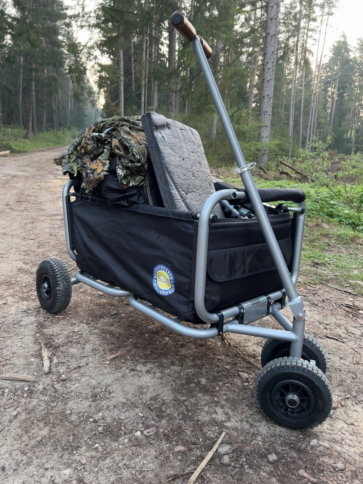 Grey garden cart with camouflage fabric and telephoto lens on a sandy forest track among conifers