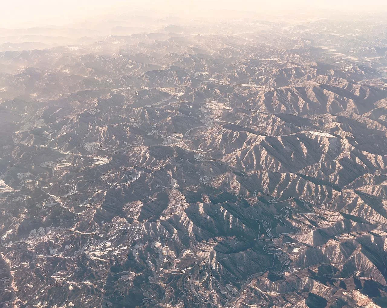 Aerial view from a plane: furrowed mountain ridges with patches of snow fading into warm morning haze towards the horizon.