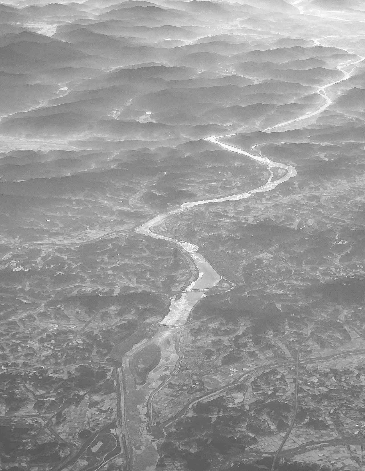 Aerial view: a river winds like a silver ribbon through hazy hill ranges dotted with fields, settlements and bridges.