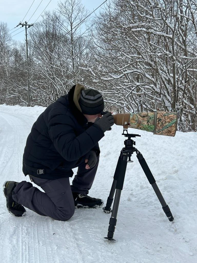 Photographe agenouillé dans la neige devant un appareil sur trépied recouvert d'un camouflage, arbres enneigés en arrière-plan.
