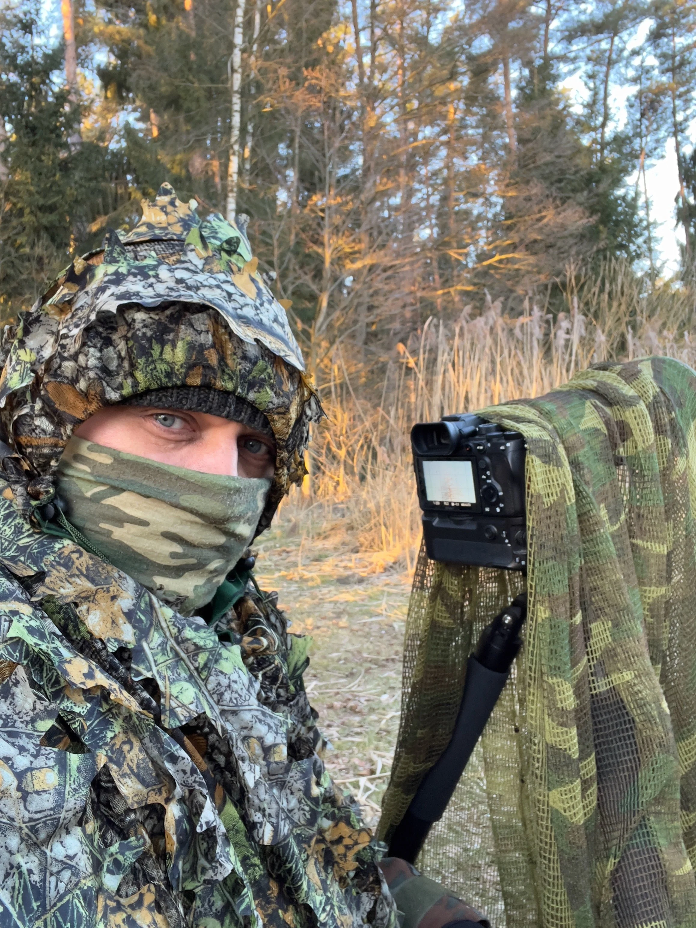A photographer from WildeNatur in camouflage clothing and face mask sits in nature next to a camouflaged camera on a tripod to photograph wildlife unnoticed.