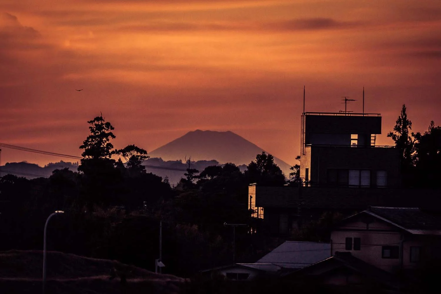 Coucher de soleil rouge orangé au-dessus de toits japonais, avec un cône montagneux lointain dans la brume