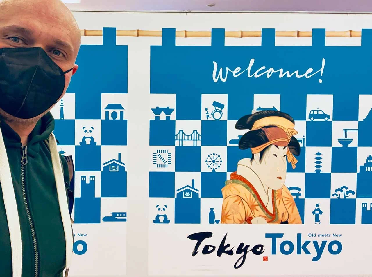 Selfie with black face mask in front of the blue-and-white "Welcome! Tokyo Tokyo" wall with ukiyo-e figure and city pictograms.