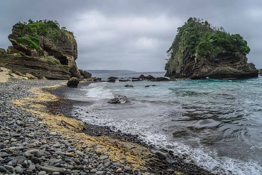 Pebble beach with shallow waves and two vegetated rock formations under an overcast sky, Izu, Japan.