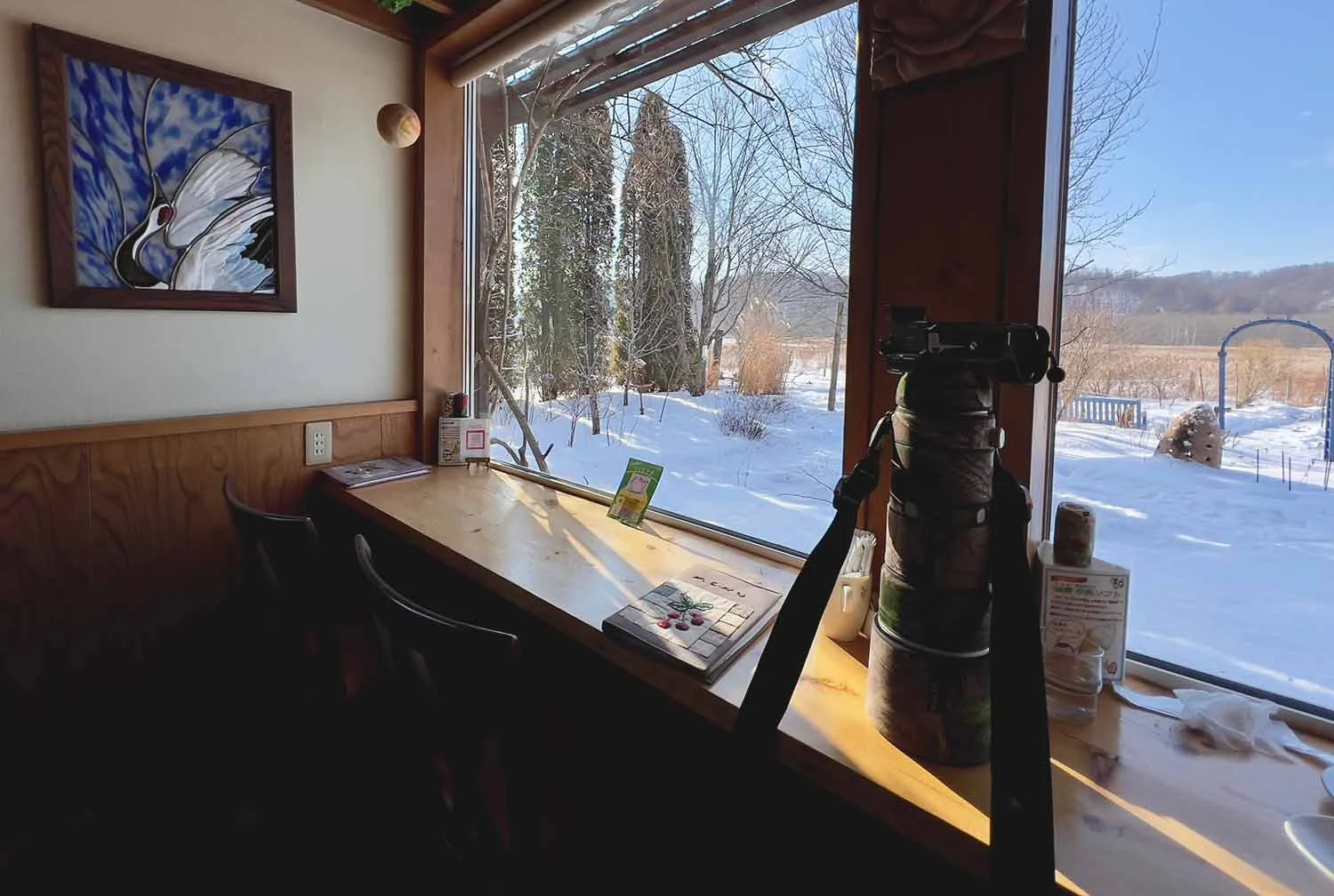 Window seat with telephoto lens on the sill, stained-glass crane picture on the wall, snowy view outside.
