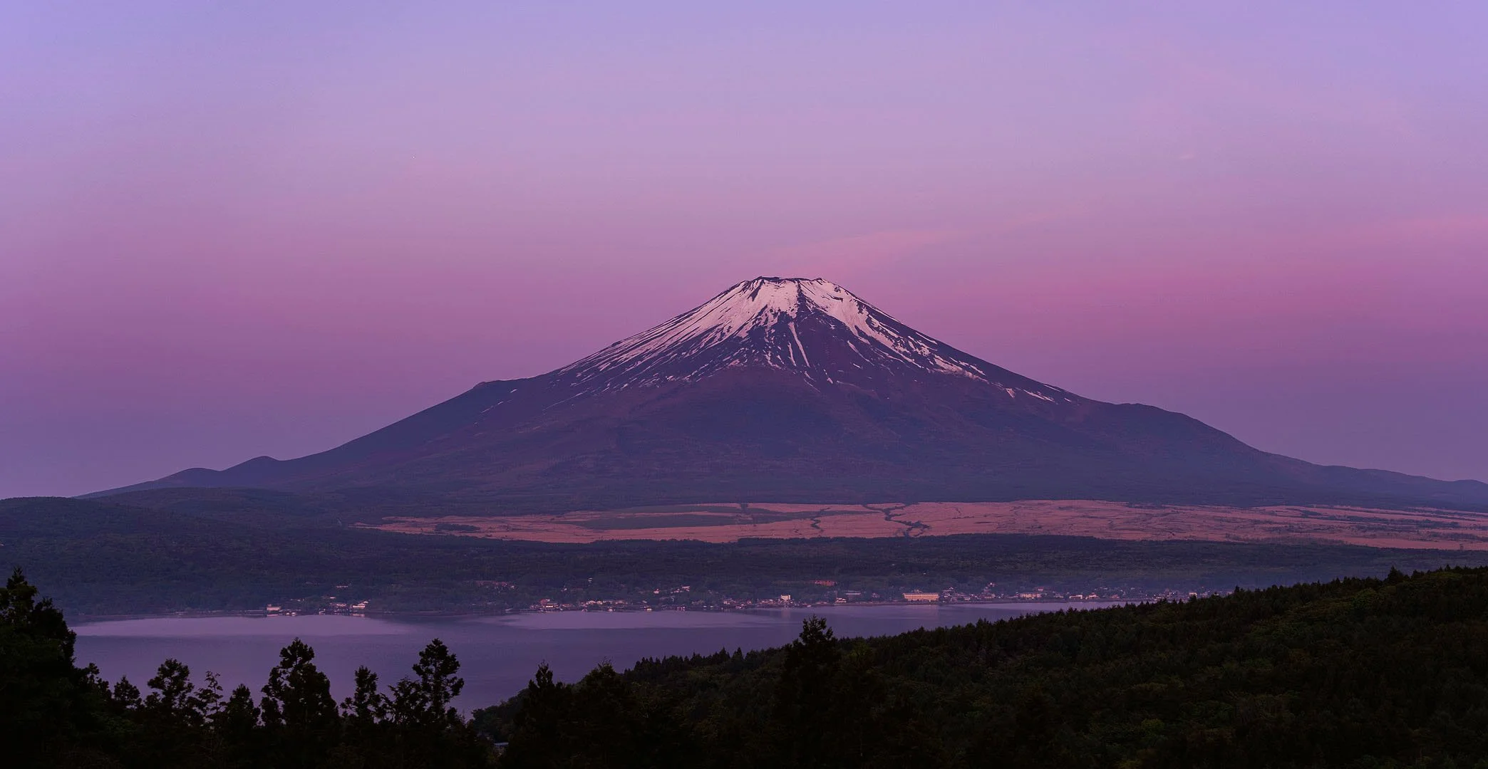 Mount Fuji with its snow-covered summit in violet evening light above a lake in Japan.