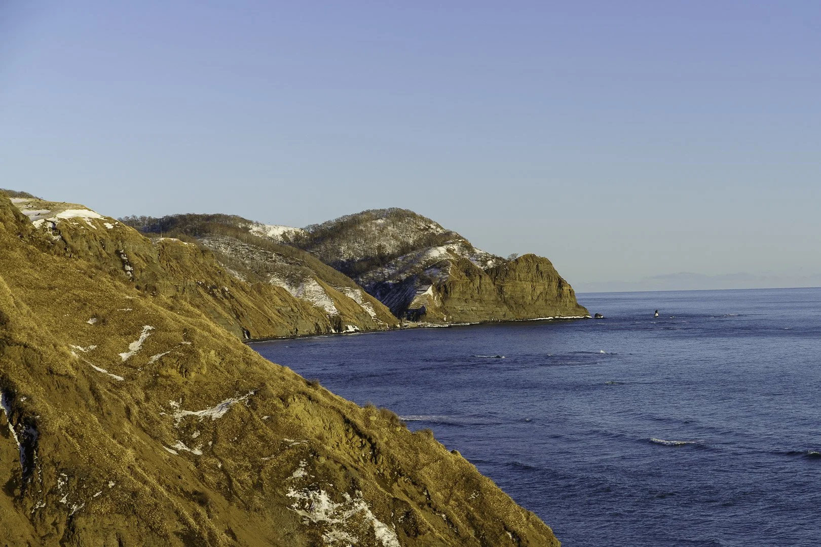 Falaises côtières abruptes striées de neige plongeant dans une mer bleu profond, derrière une ligne de collines enneigées.