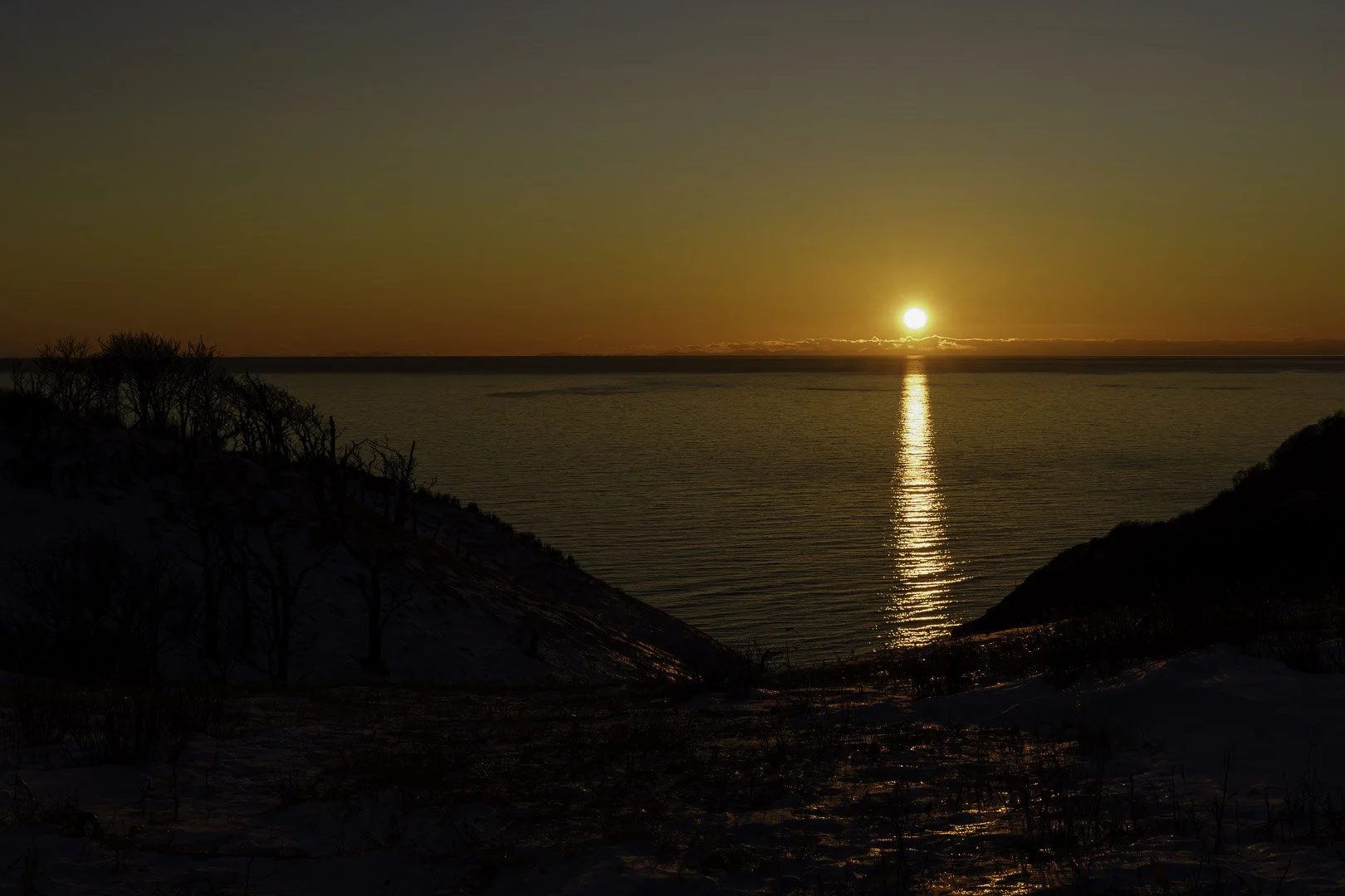 Petit disque solaire au-dessus d’une mer calme avec une traînée de lumière dorée, encadré par des versants enneigés sombres.