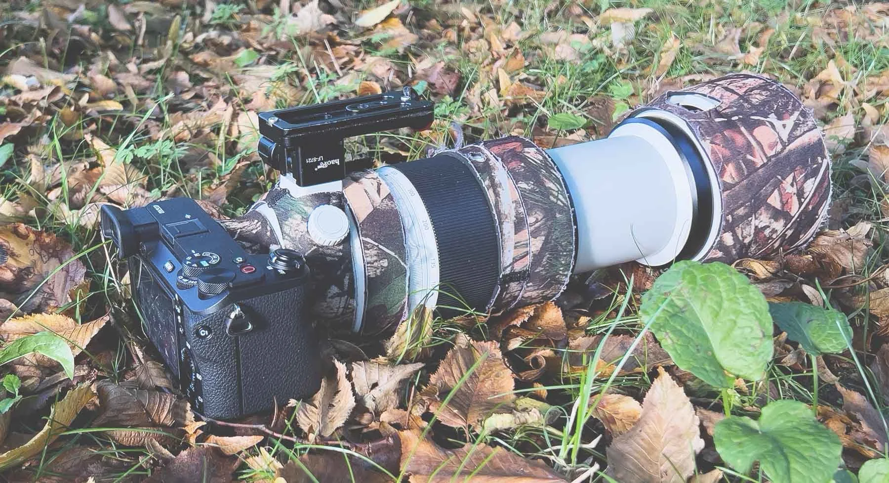 Camera with telephoto zoom lens in camouflage sleeve on forest floor, ideal for wildlife photography.