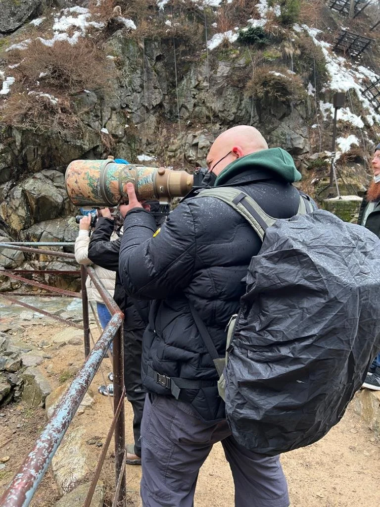 Photographer with a camouflaged super-telephoto lens above a rocky, snowy river valley.